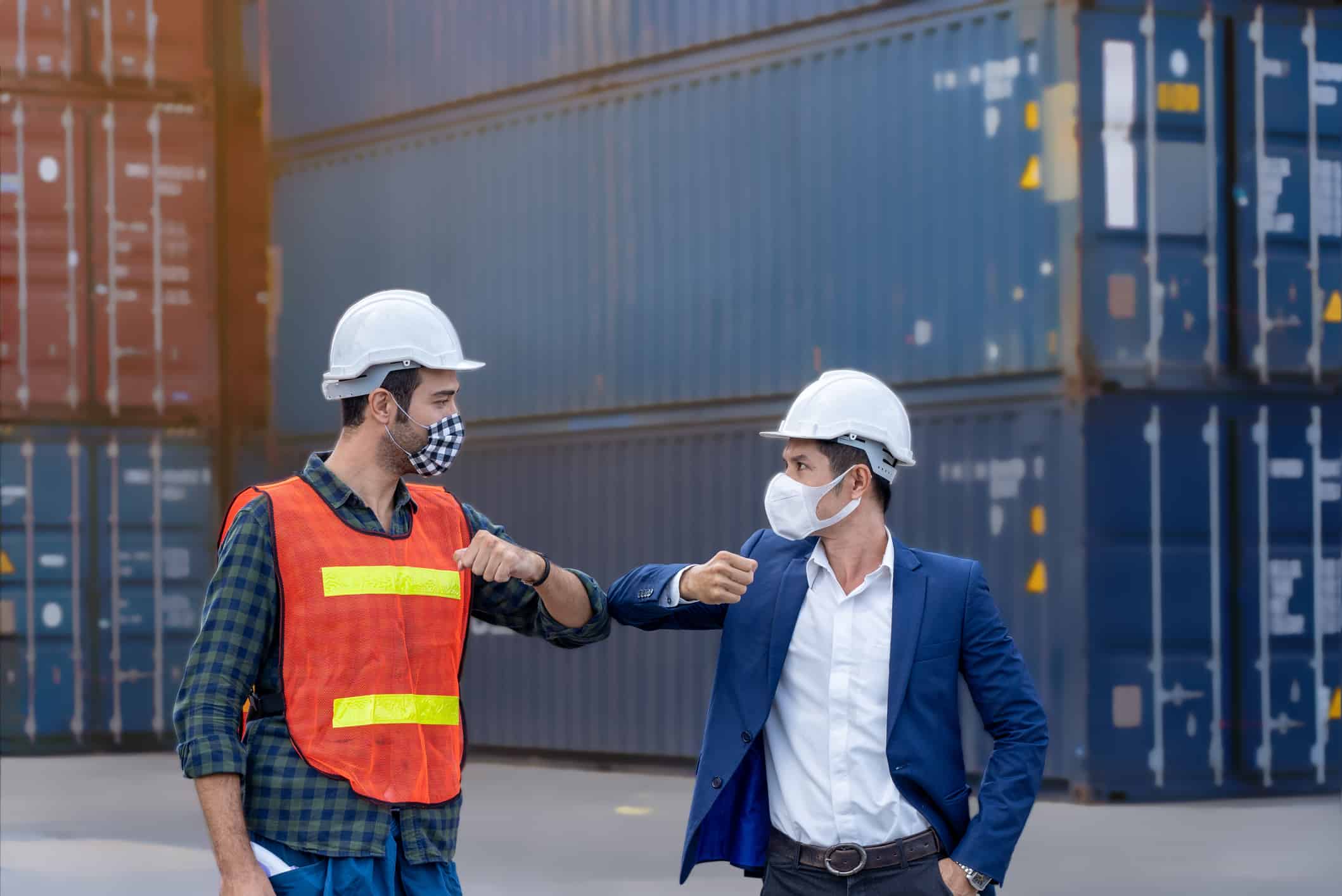 Engineer worker greeted by bumping elbow for distance between people wearing mask and helmet in the factory or container yard Prevent accident from work or dust and Coronavirus.