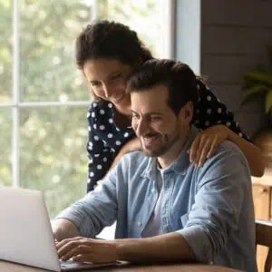 Affectionate young hispanic woman cuddling shoulders of happy man, using computer applications, choosing goods in internet store, reading email with pleasant news, typing message in social network.