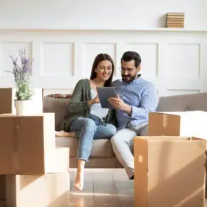 Full length happy family sitting on couch among cardboard boxes in new house, using digital tablet, buying decorations online. Smiling couple enjoying moving in new apartment, shopping in internet.